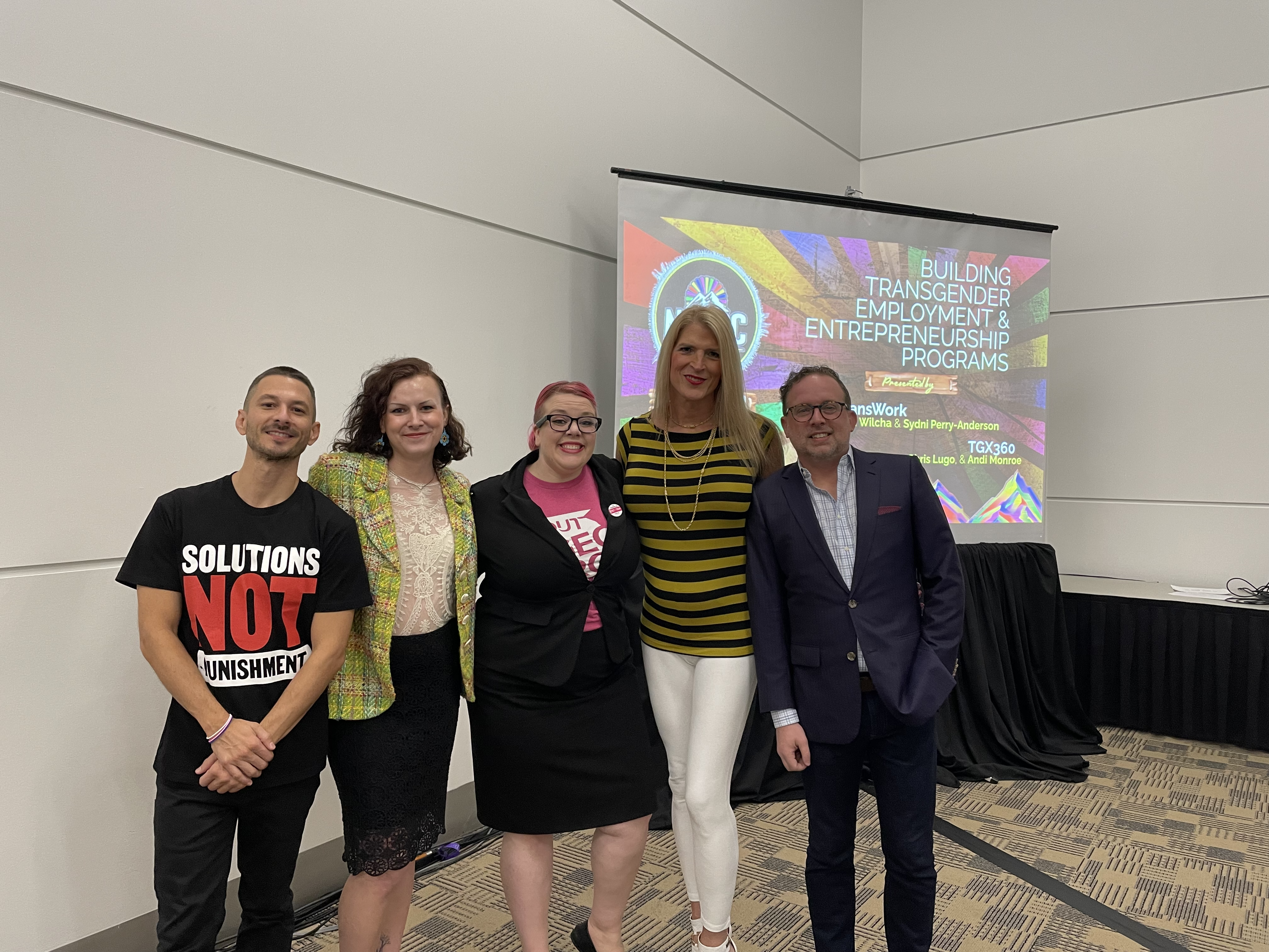 A group of five standing shoulder-to-shoulder in front of a drop-down projection screen. It is the presenters from TransWork, the IBA, OUT Georgia, and TGX360. They are dressed in business casual clothing, and on the screen there is text reading "Building Transgender Employment and Entrepreneurship Programs"