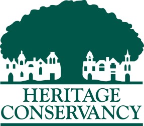 the heritage conservancy logo, green text displays the company name below a tree of the same color. an outline of buildings is in front of the tree.